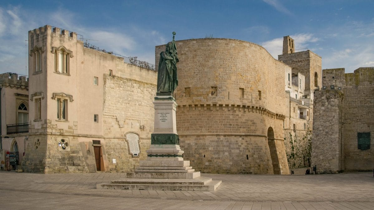 A statue stands high on a plinth in front of thick medieval walls.